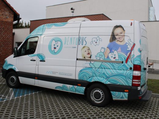 Zdjęcie dentobusu. Samochód stoi na szkolnym parkingu. Jest biały i oklejony grafikami dzieci, szczoteczki, zęba z uśmiechem i tubki pasty. Po środku widnieje napis "Dentobus".