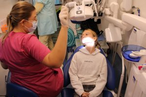 Uczennica siedzi na fotelu dentystycznym. Głowę ma zwróconą w kierunku Pani doktor siedzącej obok. Pani doktor ustawia lampę oświetlającą część twarzy dziewczynki.