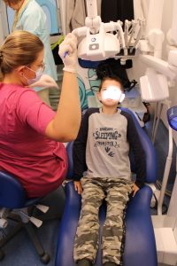 Chłopiec siedzi na fotelu dentystycznym. Jego twarz oświetlona jest lampą, która ustawia siedząca obok Pani doktor.