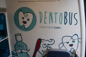 Zdjęcie jednej ze ścian w dentobusie. Na białym tle grafiki przedstawiające uśmiechnięte zęby, szczoteczkę i płyn do dezynfekcji jamy ustnej. po środku napis "DENTOBUS I NIESFORNE ZĄBKI".