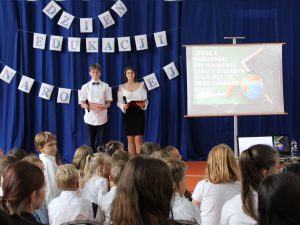 Na niebieskim tle z zawieszonym napisem "DZIEŃ EDUKACJI NARODOWEJ" stoi dwoje uczniów prowadzących akademię. Obok nich ustawiony jest stojak z białym ekranem, na którym wyświetla się tekst z rzutnika: "JEDNĄ Z NAJBARDZIEJ DECYDUJĄCYCH RZECZY W NASZYM ŻYCIU JEST TO, GDZIE CHODZIMY DO SZKOŁY". U dołu zdjęcia widać głowy siedzących na przeciwko uczniów.