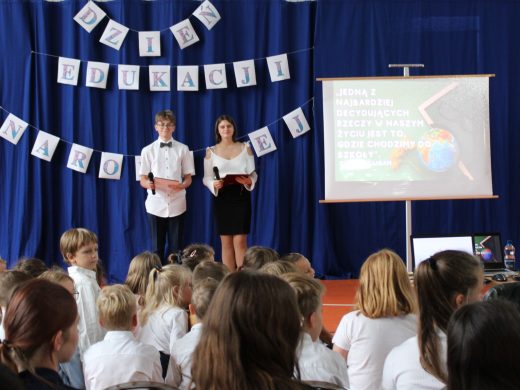 Na niebieskim tle z zawieszonym napisem "DZIEŃ EDUKACJI NARODOWEJ" stoi dwoje uczniów prowadzących akademię. Obok nich ustawiony jest stojak z białym ekranem, na którym wyświetla się tekst z rzutnika: "JEDNĄ Z NAJBARDZIEJ DECYDUJĄCYCH RZECZY W NASZYM ŻYCIU JEST TO, GDZIE CHODZIMY DO SZKOŁY". U dołu zdjęcia widać głowy siedzących na przeciwko uczniów.