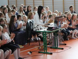 Zdjęcie uczniów siedzących na widowni. Centralnie po środku siedzi za małym stolikiem nauczycielka przy laptopie trzymająca w dłoni mikrofon.