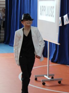 Uczeń w stroju Michaela Jacksona schodzi ze sceny. Podnosi lekkow górę głowę a jego twarz częściowo zasłonięta jest przez czarny kapelusz. Obok niego na rzutniku wyświetla się napis " APPLAUSE".