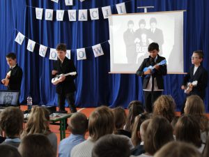 Czworo uczniów stoi na scenie z kolorowymi ukulele. Chłopcy odtwarzają fragment występu grupy the beatles. Za nimi widać granatowe tło i rzutnik na którym wyświetla się jedna z okładek zespołu. Przed chłopcami widać głowy widowni.