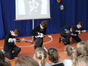 Czworo uczniów klęka na jednym kolanie na scenie z kolorowymi ukulele. Chłopcy odtwarzają fragment występu grupy the beatles. Za nimi widać granatowe tło i rzutnik na którym wyświetla się jedna z okładek zespołu. Przed chłopcami widać głowy widowni.