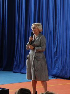 Pani Dyrektor podczas wystąpienia na akademii. W ręku trzyma mikrofon i się uśmiecha. W tle widać granatowy materiał.