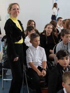 Nauczycielka matematyki wyprostowana i uśmiechnięta stoi przy swoich uczniach siedzących podczas akademii na krzesełkach.