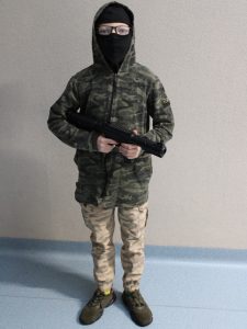 Uczeń w stroju żołnierza stoi na szkolnym korytarzu. W dłoni ma zabawkowy karabin a na twarzy kominiarkę.
