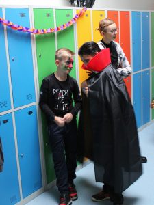 Troje uczniów stoi przy kolorowych szkolnych szafkach. Uczeń przebrany za wampira pochyla się do swojego kolegi, który ma pomalowaną na kolorowo twarz.