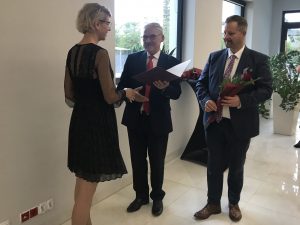 Wójt gminy Łysomice wraz z przewodniczącym Rady gminy wręczają nauczycielce stojącej bokiem do zdjęcia nagrodę. Wójt trzyma dyplom umieszczony w sztywnej oprawie a przewodniczący kwiaty.