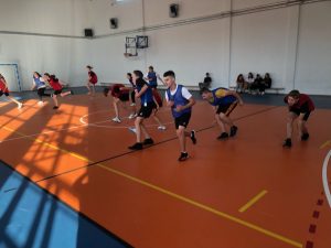 Uczniowie biegną przez salę gimnastyczną. Część z nich schyla się dotykając dłońmi parkietu. Zdjęcie wykonane w ruchu.