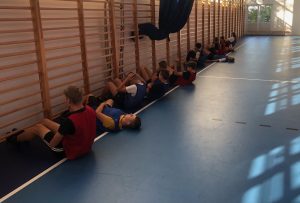 Uczniowie leżą na plecach przy drabinkach na sali gimnastycznej. Część dzieci podnosi się robiąc skłony.