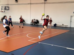 Zdjęcie wykonane w ruchu. Chłopiec na pierwszym planie trzyma w dłoni piłkę z którą wyskakuje w górę. W tle widać innych biegnących uczniów.