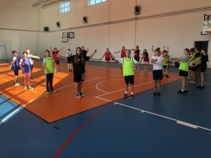 Uczniowie stoją na sali gimnastycznej po okręgu. Podnoszą do góry ręce, którymi wymachują. Część dzieci zwrócona jest przodem do zdjęcia.