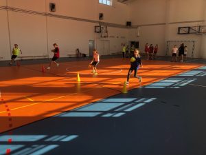 Czworo uczniów biegnie kozłując piłkami. Przed uczniami stoją pachołki w równych odstępach do których uczniowie się zbliżają.