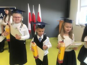 Troje uczniów klasy pierwszej stoi obok siebie. Dzieci ubrane są na galowo a na głowach maja papierowe birety. W dłoniach trzymają pudełka w kształcie kredek, dyplomy oraz książki.
