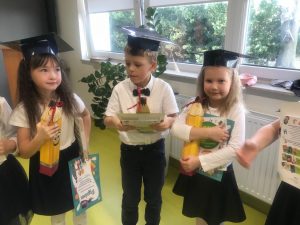 Troje uczniów z klasy pierwszej stoi obok siebie w galowych strojach. Dzieci na głowie mają papierowe birety a w dłoniach trzymają pudełka w kształcie ołówków, książki i dyplomy.