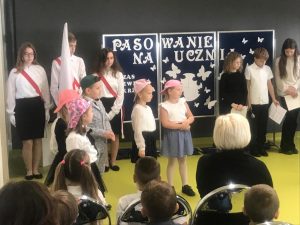 Przy granatowej tablicy z napisem "PASOWANIE NA UCZNIA" stoi trzech uczniów z głowami spuszczonymi na dół oraz poczet flagowy szkoły. Przed nimi stoi piątka uczniów klasy pierwszej w kolorowych czapeczkach z daszkiem na głowie.