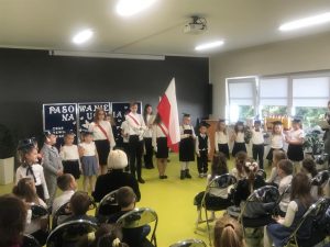 Uczniowie pierwszej klasy stoją w półokręgu. Po środku stoi poczet flagowy szkoły z opuszczoną w przód flagą. Przed uczniami na krzesłach siedzi widownia.