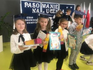 Czworo uczniów klasy pierwszej stoi obok siebie. Dzieci ubrane są na galowo a na głowach mają papierowe birety. W dłoniach trzymają pudełka w kształcie kredek, dyplomy oraz książki. Dzieci oglądają otrzymane upominki. W tle widać granatową tablicę z białym napisem "PASOWANIE NA UCZNIA".