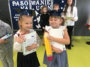 Dwie uczennice klasy pierwszej stoją obok siebie. Dzieci ubrane są na galowo a na głowach maja papierowe birety. W dłoniach trzymają pudełka w kształcie kredek, dyplomy oraz książki. Dziewczynki się uśmiechają. Za nimi widać granatową tablicę z białą dekoracją. oraz troje uczniów klasy czwartej stojących przed nią.