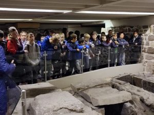 Uczniowie stoją wzdłuż przezroczystej balustrady i oglądają zamieszczone za nią kamienie odkryte w piwnicach katedry gnieźnieńskiej.