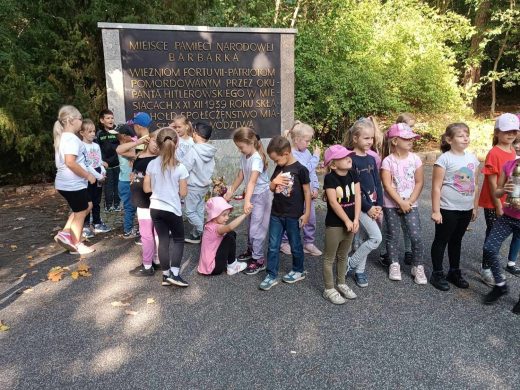 Uczniowie stoją w rzędzie odwróceni plecami do pomnika z napisem "MIEJSCE PAMIĘCI NARODOWEJ BARBARKA".