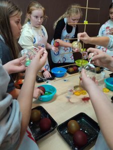 Uczniowie stoją przy stoliku i dekorują swoje bombki. Na stole po środku ustawiony jest stojak na bombki. Dzieci maja na sobie ochronne fartuchy.