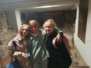 Trzy dziewczynki pozują do wspólnego zdjęcia w piwnicy katedry gnieźnieńskiej. Dziewczynki się uśmiechają. W tle widać zachowane elementy wcześniejszej świątyni.