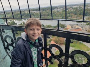 Chłopiec pozuje do zdjęcia na punkcie widokowym. Za nim widać zdobioną balustradę oraz płynąca przez Gniezno rzekę.