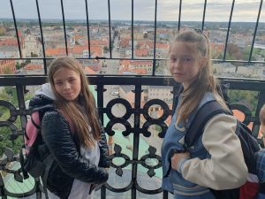 Dwie dziewczynki pozują do zdjęcia na punkcie widokowym. Za nimi widać metalową, zdobioną balustradę oraz zabudowania miasta Gniezno.