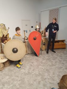 Dwóch chłopców pozuje do zdjęcia w strojach wojów,. Obaj trzymają duże tarcze - jeden okrągłą drugi czerwona w kształcie łzy oparta o podłogę. Obok stoi Pan prowadzący zajęcia i wskazuje na jednego z chłopców.