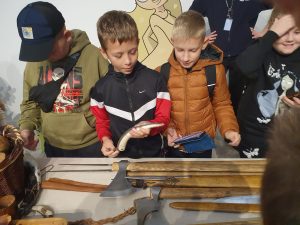 Trzech uczniów stoi przy stole, na którym leżą topory. jeden z nich trzyma w dłoni wygiętą w łuk broń sieczną.