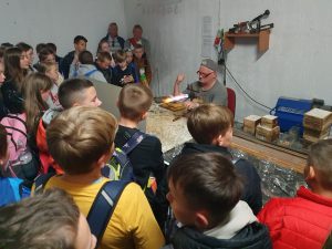 Uczniowie stoją zgromadzenie przy stole za którym siedzi Pan rozgrzewający włączonym palnikiem szklaną rurkę. Uczniowie obserwują jego pracę.