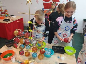 Dwoje uczniów stoi przy stoliku trzymając w dłoniach własnoręcznie ozdabiane bombki. Dzieci mają na sobie białe fartuszki ozdobione kołami, gwiazdami i piorunami. Na stole przy którym pracują ustawiony jest wieszak na bombki częściowo już zapełniony. W tle widać innych pracujących uczniów.