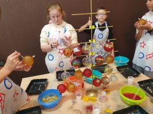 Uczniowie stoją przy stole i dekorują bombki. Na stole przed nimi stoi duży stojak z zawieszonymi gotowymi bombkami. Zza stojaka widać pracującą uczennicę i ucznia trzymającego podniesiony w górę kciuk.