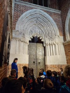 Zdjęcie drzwi gnieźnieńskich obok których stoi Pani przewodnik. na przeciwko widać stojących uczniów.