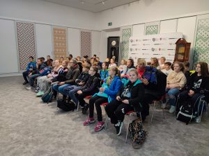 Uczniowie siedzą na krzesełkach w dużej sali i słuchają rozpoczynającej się lekcji muzealnej o historii powstania państwa polskiego.