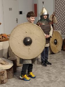 Dwóch uczniów stoi w przebraniach wojów. Jeden z nich trzyma ogromną tarczę i się uśmiecha.