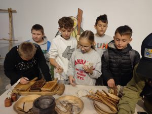 Uczniowie stoją przy stole na którym ustawione są repliki narzędzi i przedmiotów użytku codziennego z dawnych czasów. Uczniowie przyglądają się zgromadzonym przedmiotom.