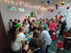 Uczniowie podczas warsztatów z dekorowania bombek. dzieci stoją przy kilku stołach. Na ścianach sali widać motywy świąteczne a nad sufitem zawieszone są kolorowe bombki i lampki choinkowe.