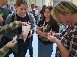 Nauczycielka wypełnia karteczkę z zadaniami z tabliczki mnożenia. Stoi bokiem a przed nią uczniowie trzymający kopertę.