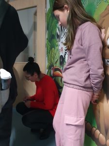 Nauczycielka kuca przy ścianie rozwiązując zadania z tabliczki mnożenia. Obok stoi oparta o ścianę uczennica.