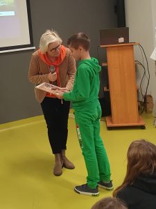 Pani Dyrektor wręcza nagrodę uczniowi. Chłopiec trzyma w dłoniach książkę, na którą patrzy, a Pani Dyrektor pochyla się do niego.