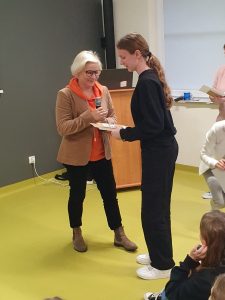 Pani Dyrektor wręcza nagrodę uczennicy. Dziewczynka trzyma w dłoniach dyplom i książkę na które patrzy.