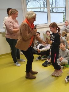 Pani Dyrektor wręcza nagrodę uczennicy. W tle widać siedzących na podłodze uczniów.