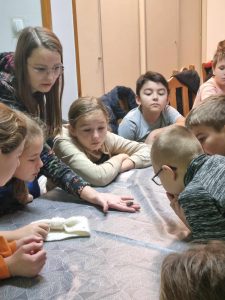 Uczniowie siedzą przy dużym stole i pochylają się przed siebie. Obserwują przedmiot umieszczony na dłoni Pani prowadzącej warsztaty. Pani trzyma dłoń opartą o stół.