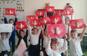 Uczniowie oddziału przedszkolnego pozują do wspólnego zdjęcia. Dzieci wyciągają wysoko w górę ręce ze swoimi praca, na których jest orzełek.