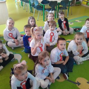 Uczniowie oddziału przedszkolnego siedzą na dywanie. Ubrani są w stroje galowe, a do bluzek mają przypięte biało czerwone kotyliony.